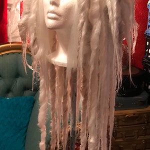 Puede incluir: Una peluca blanca con rastas largas y gruesas. La peluca est&aacute; peinada en una cabeza de maniqu&iacute; y se muestra en un soporte.