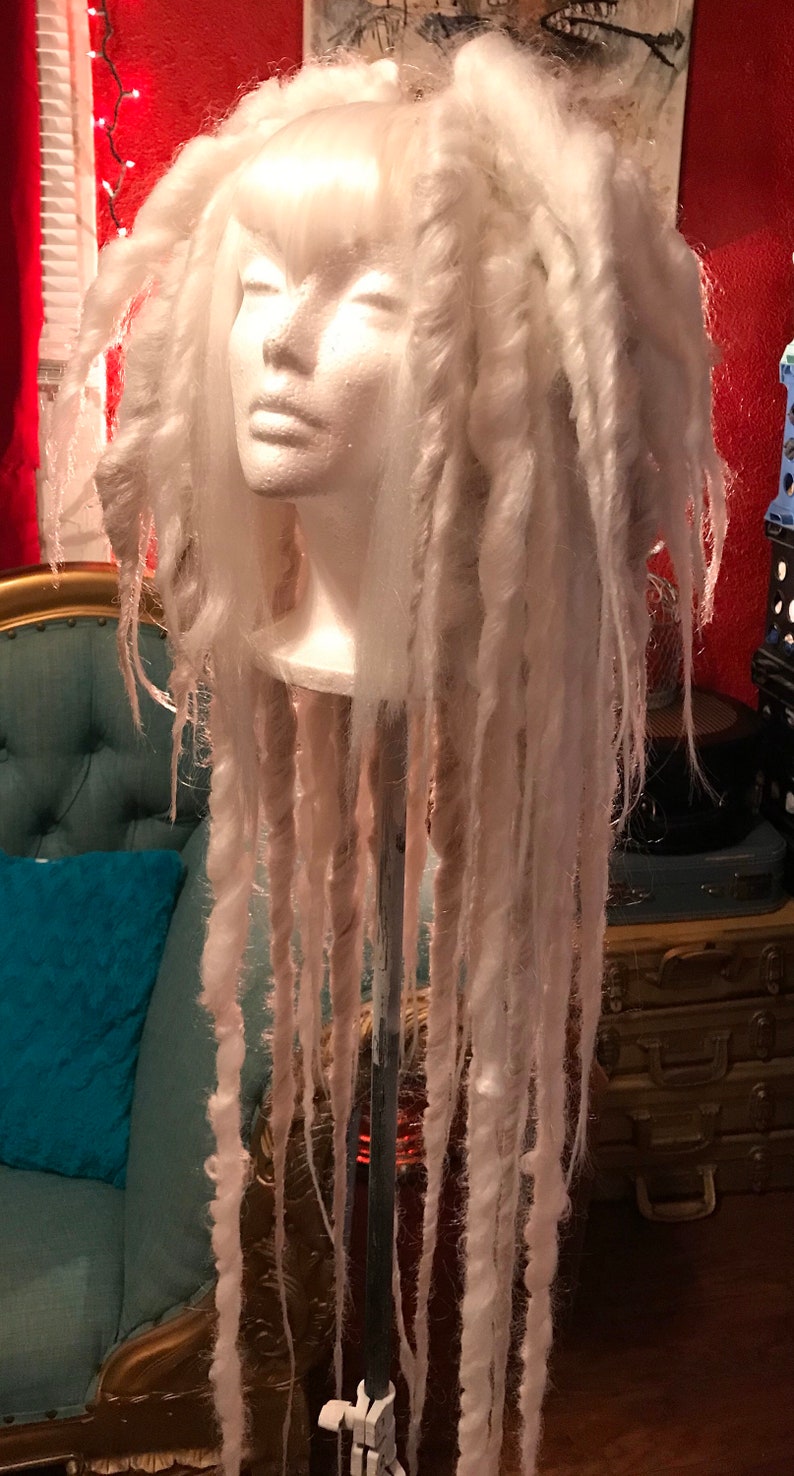 Puede incluir: Una peluca blanca con rastas largas y gruesas. La peluca est&aacute; peinada en una cabeza de maniqu&iacute; y se muestra en un soporte.