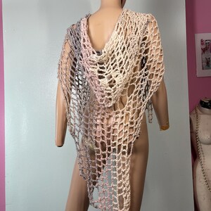 May include: A handmade, open-weave crochet shawl in a gradient of colors, including gray, pink, and beige. The shawl features a hood and drapes over the shoulders. The garment is displayed on a mannequin.