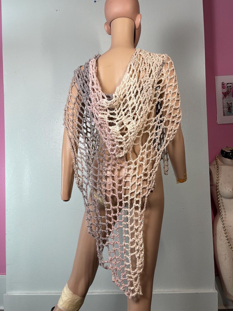 May include: A handmade, open-weave crochet shawl in a gradient of colors, including gray, pink, and beige. The shawl features a hood and drapes over the shoulders. The garment is displayed on a mannequin.