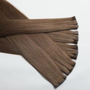 Puede incluir: Varios paquetes de extensiones de cabello castaño claro se colocan sobre un fondo blanco. Las extensiones son lisas y parecen estar hechas de cabello humano. Los paquetes están cosidos en la parte superior, creando una trama.