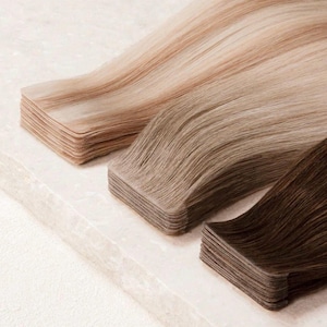 Puede incluir: Tres paquetes de extensiones de cabello en tonos rubios, castaños claros y castaños oscuros. El cabello está en capas y parece suave y brillante. Las extensiones están cuidadosamente apiladas, mostrando las diferentes opciones de color.