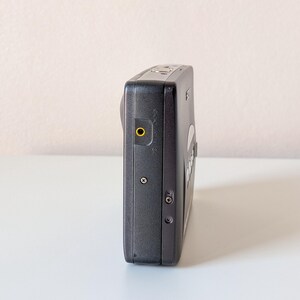 May include: A black digital camera viewed from the side, featuring a prominent yellow-rimmed circular port, two small exposed screws on the side panel, a metallic shutter button on the top edge, and a Sony logo visible on the upper corner.
