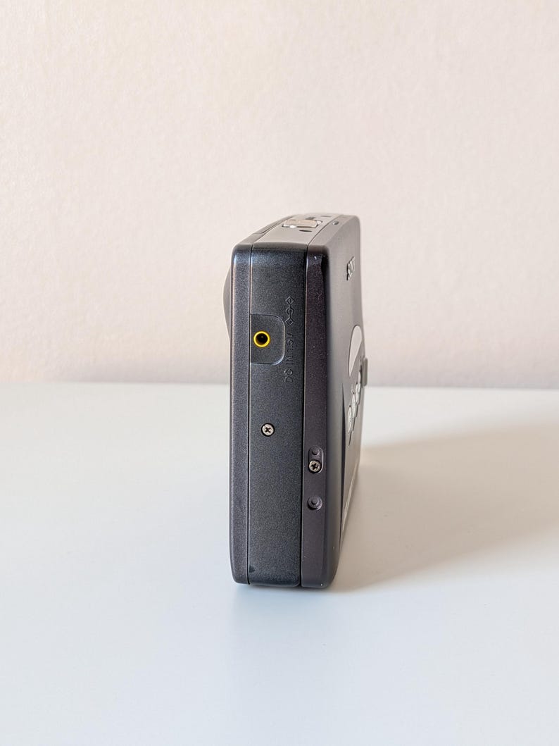 May include: A black digital camera viewed from the side, featuring a prominent yellow-rimmed circular port, two small exposed screws on the side panel, a metallic shutter button on the top edge, and a Sony logo visible on the upper corner.