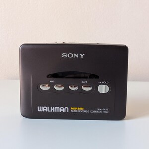 May include: A black Sony Walkman cassette player featuring the model number WM-EX53, Mega Bass, and Auto Reverse technology. The device includes silver control buttons labeled AMS, BATT, and a white sliding HOLD switch on the front panel.