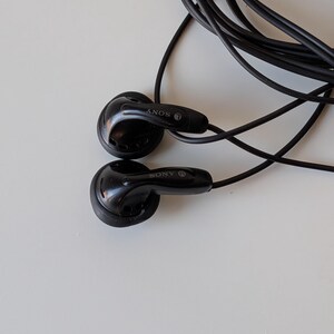 May include: Black wired earbuds with a long, thin cable. Each earbud features a glossy finish and the Sony brand name printed in white. The left earbud is marked with an L and the right earbud is marked with an R. The cord is coiled behind the buds.