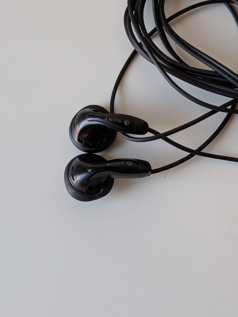 May include: Black wired earbuds with a long, thin cable. Each earbud features a glossy finish and the Sony brand name printed in white. The left earbud is marked with an L and the right earbud is marked with an R. The cord is coiled behind the buds.