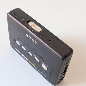 May include: A dark gray Sony Walkman portable cassette player featuring a top-mounted silver open latch, four circular control buttons, and a white hold switch. The front casing displays the Sony logo, Walkman branding, and Mega Bass text.