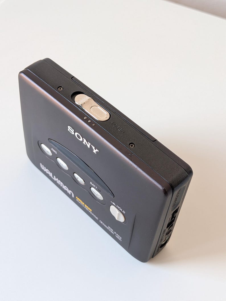 May include: A dark gray Sony Walkman portable cassette player featuring a top-mounted silver open latch, four circular control buttons, and a white hold switch. The front casing displays the Sony logo, Walkman branding, and Mega Bass text.