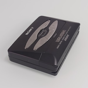 May include: Black Sony Walkman cassette player with silver controls. The device features the word "SONY" on the top left and "WALKMAN" on the bottom right. The model is WM-EX570. It has buttons for rewind, fast forward, and other functions.