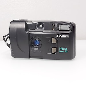 Può includere: Fotocamera a pellicola Canon Prima Junior DX nera. La fotocamera ha un flash integrato, un obiettivo e un cinturino da polso. Il logo Canon è bianco e il testo Prima Junior DX è turchese. La fotocamera è su una superficie bianca.