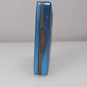 以下が含まれることがあります： 青とグレーのポータブルCDプレーヤー。デバイスは、丸みを帯びたエッジを持つ長方形の形状をしています。側面図には、「OPEN」と書かれた灰色のボタンと小さな銀色のネジが表示されています。デバイスは白い表面にあります。