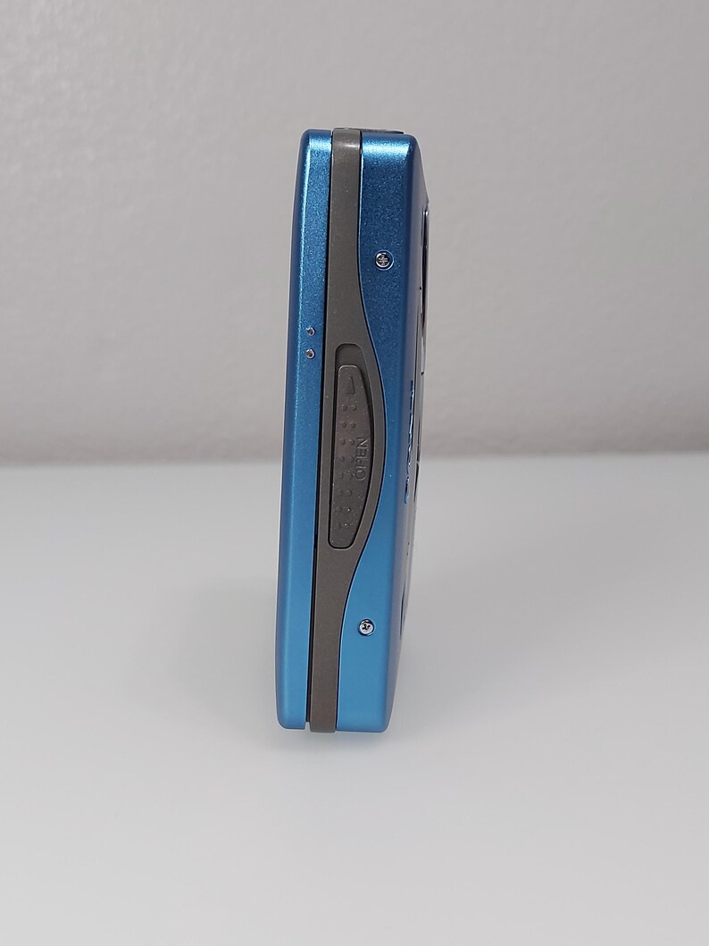 以下が含まれることがあります： 青とグレーのポータブルCDプレーヤー。デバイスは、丸みを帯びたエッジを持つ長方形の形状をしています。側面図には、「OPEN」と書かれた灰色のボタンと小さな銀色のネジが表示されています。デバイスは白い表面にあります。
