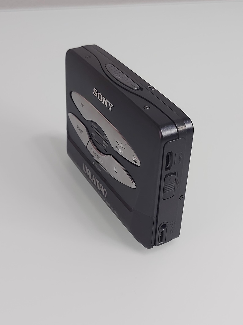 May include: Black Sony Walkman cassette player with silver buttons. The device has the word "SONY" printed on the top and "WALKMAN" on the bottom. The buttons include "FF", "REW", "FUNCTION", "REPEAT", and "BATT".