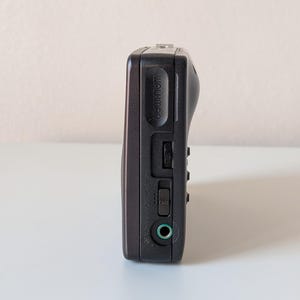 May include: A dark gray portable cassette player featuring the word Walkman embossed on the side. The device includes a volume dial, a sliding power switch labeled OFF and PHONES, and a circular green-rimmed headphone jack port on the side panel.