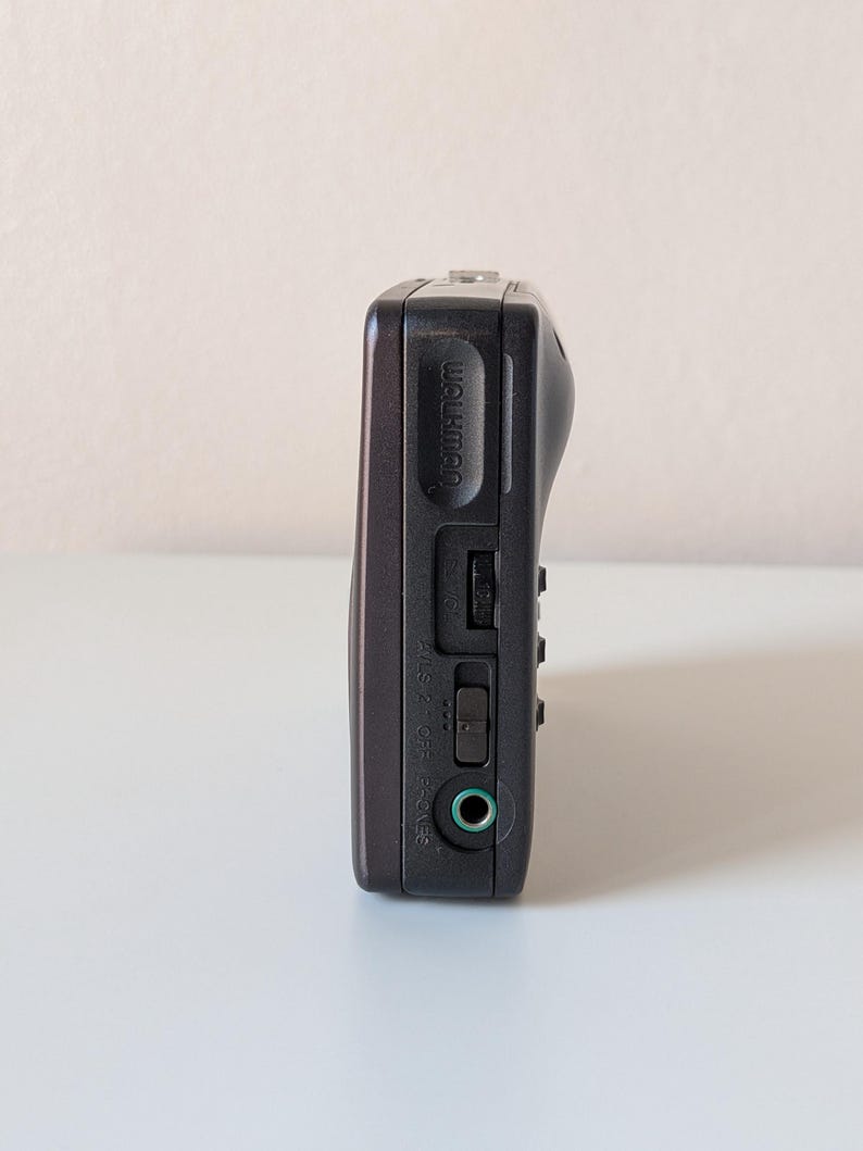 May include: A dark gray portable cassette player featuring the word Walkman embossed on the side. The device includes a volume dial, a sliding power switch labeled OFF and PHONES, and a circular green-rimmed headphone jack port on the side panel.
