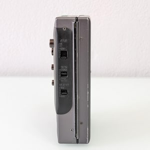 May include: A grey vintage cassette player, seen from the side, with buttons and switches. The device has a dark grey panel with labels such as "AVLS" and "TAPE". The body is a lighter grey with some wear.