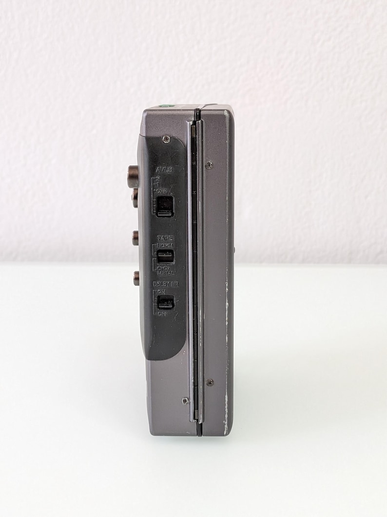May include: A grey vintage cassette player, seen from the side, with buttons and switches. The device has a dark grey panel with labels such as "AVLS" and "TAPE". The body is a lighter grey with some wear.