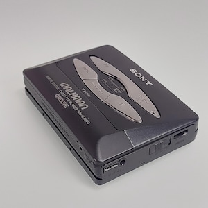 May include: A black Sony Walkman cassette player, model WM-EX570, with silver control buttons. The device has the word "GROOVE" printed on it. It features a headphone jack and a USB port. The Walkman is on a light grey background.