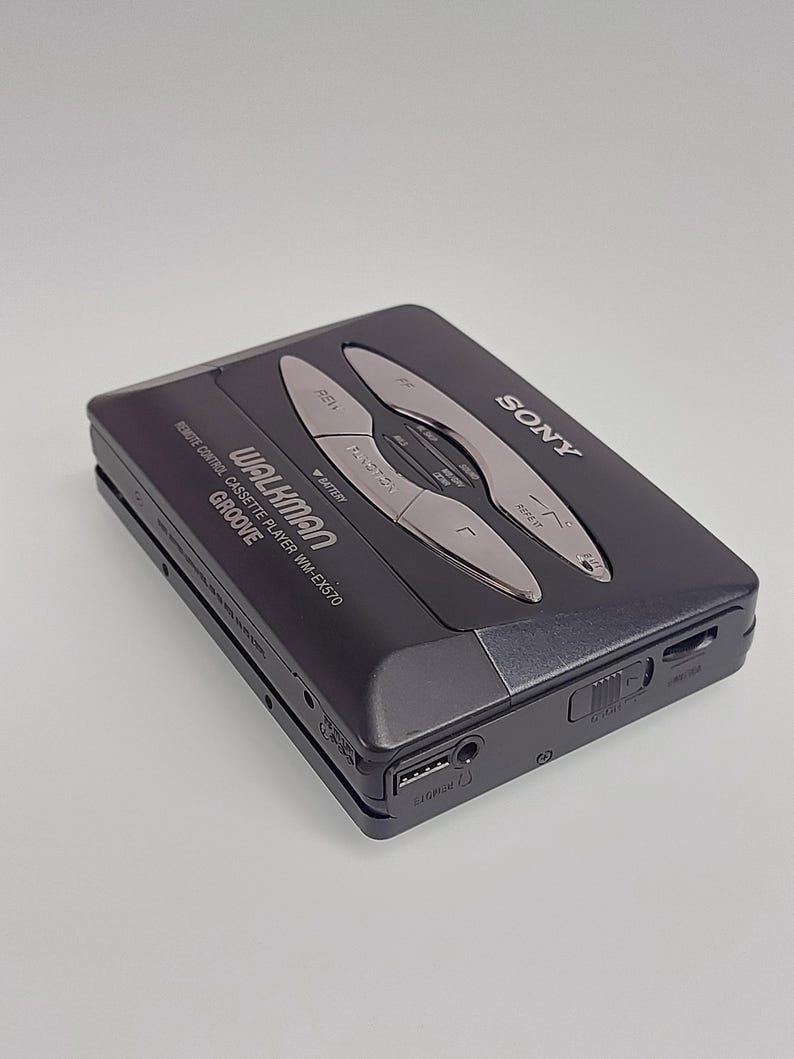 May include: A black Sony Walkman cassette player, model WM-EX570, with silver control buttons. The device has the word "GROOVE" printed on it. It features a headphone jack and a USB port. The Walkman is on a light grey background.