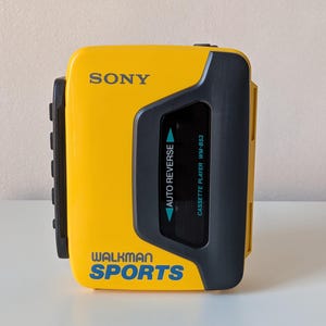 May include: A yellow Sony Walkman Sports cassette player with a dark gray plastic casing. The front features the text SONY, WALKMAN SPORTS, AUTO REVERSE, and CASSETTE PLAYER WM-B53 in blue and white lettering. It has a rectangular viewing window.