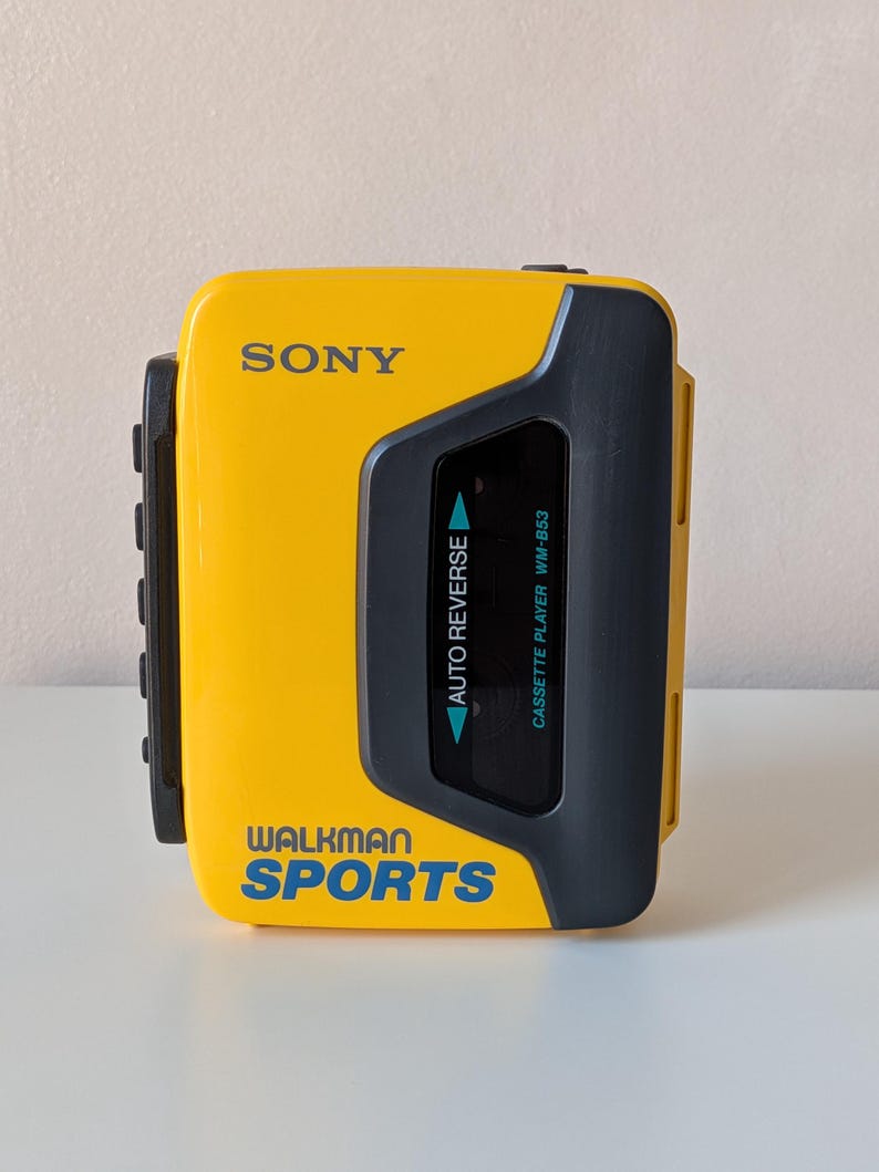 May include: A yellow Sony Walkman Sports cassette player with a dark gray plastic casing. The front features the text SONY, WALKMAN SPORTS, AUTO REVERSE, and CASSETTE PLAYER WM-B53 in blue and white lettering. It has a rectangular viewing window.