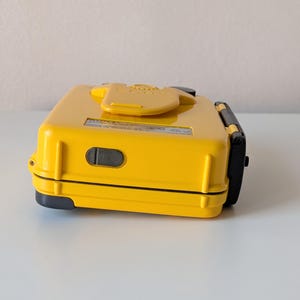 May include: A bright yellow, rectangular, hard-shell case with a black latch on the side and a central yellow clip on the top. A small black rectangular port is visible on the side, and a white label with black text is affixed to the top surface.