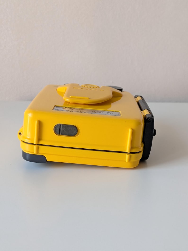 May include: A bright yellow, rectangular, hard-shell case with a black latch on the side and a central yellow clip on the top. A small black rectangular port is visible on the side, and a white label with black text is affixed to the top surface.