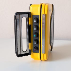 May include: A bright yellow Sony Walkman portable cassette player with its door open. The device features a black control panel with five circular buttons marked with playback symbols, including a square, triangles, and a red arrow, and a metal frame.