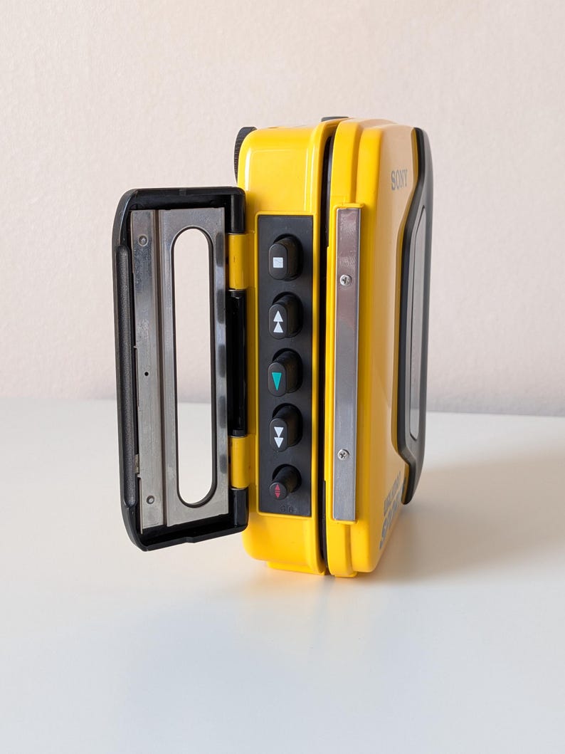 May include: A bright yellow Sony Walkman portable cassette player with its door open. The device features a black control panel with five circular buttons marked with playback symbols, including a square, triangles, and a red arrow, and a metal frame.