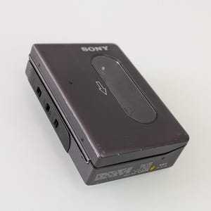 May include: A dark grey Sony Walkman cassette player. The portable audio device has a rectangular shape with a rounded top and a black control panel. The Sony logo is visible on the top. The device has a small, oval-shaped window and a directional arrow.