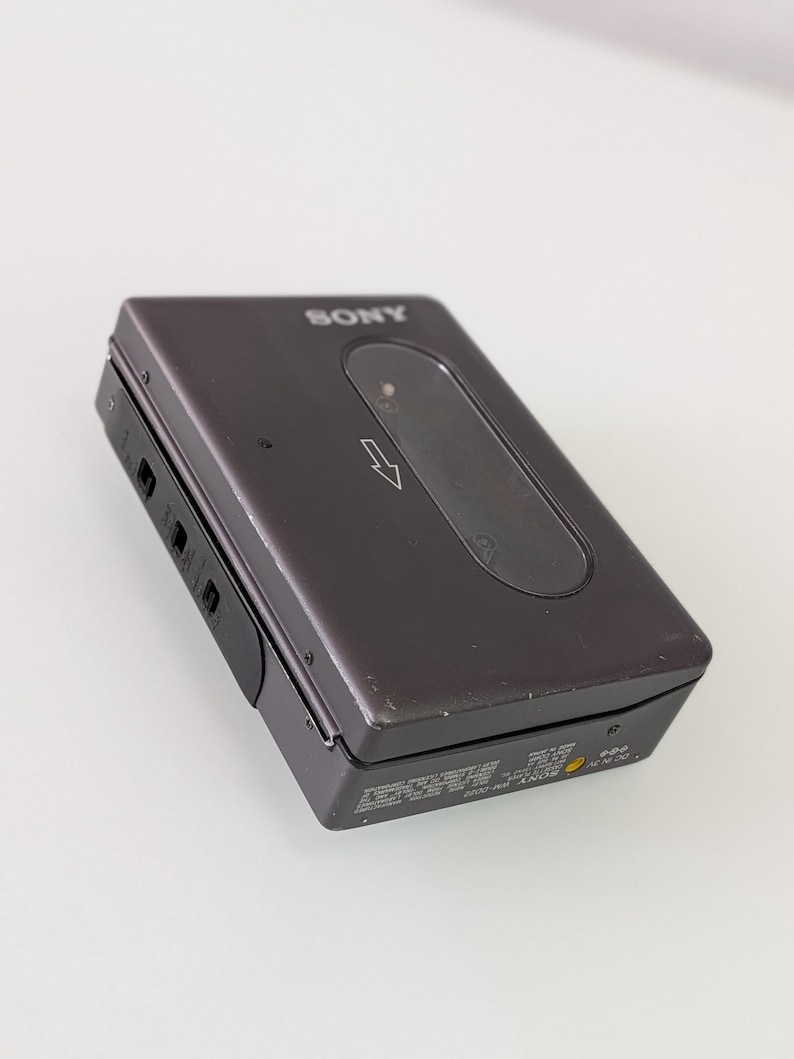 May include: A dark grey Sony Walkman cassette player. The portable audio device has a rectangular shape with a rounded top and a black control panel. The Sony logo is visible on the top. The device has a small, oval-shaped window and a directional arrow.