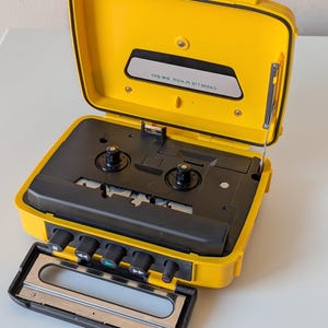 May include: A bright yellow portable cassette player with a black interior mechanism and control buttons. The lid is open, revealing a black panel with the text CASSETTE PLAYER MW-823. The device features a hinged handle and a metal antenna.