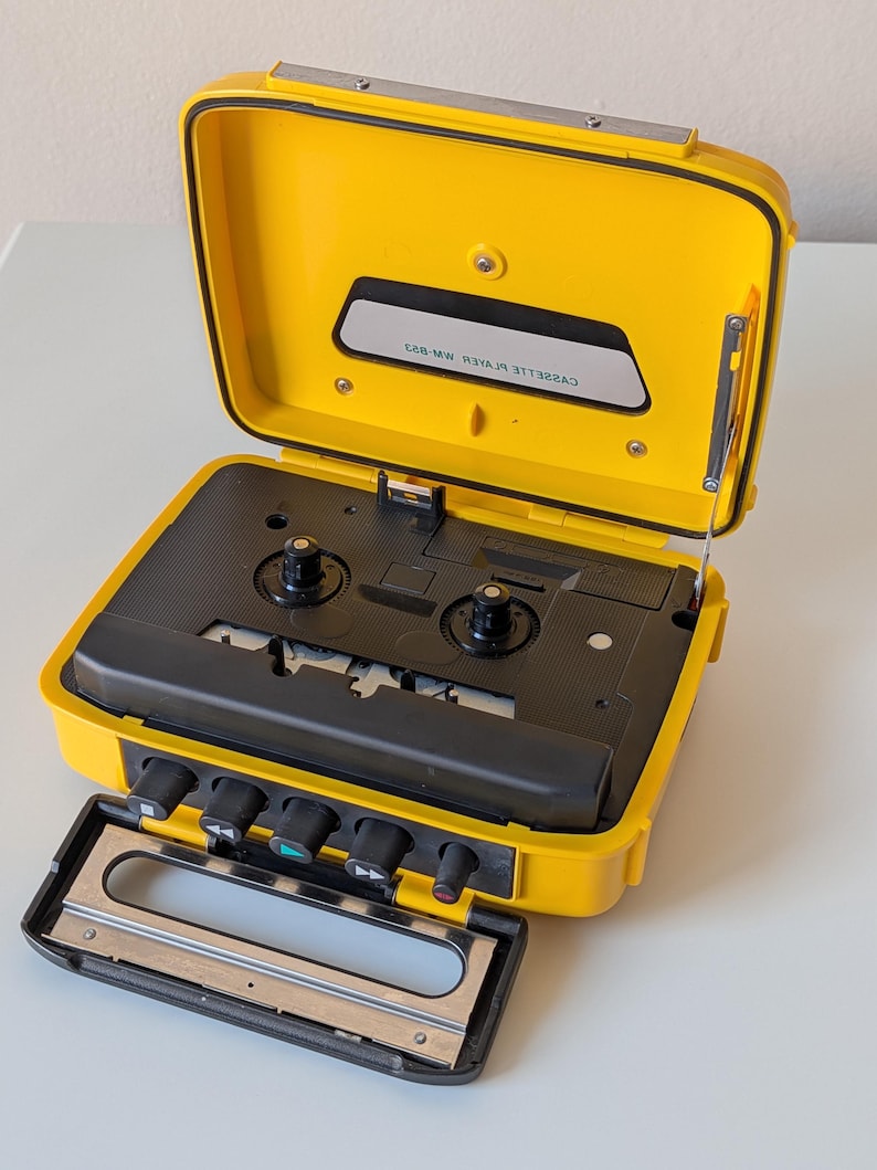 May include: A bright yellow portable cassette player with a black interior mechanism and control buttons. The lid is open, revealing a black panel with the text CASSETTE PLAYER MW-823. The device features a hinged handle and a metal antenna.