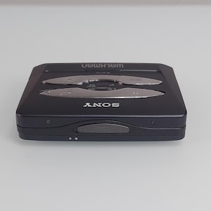 May include: A black Sony Walkman cassette player. The device features silver buttons for rewind, fast forward, and play. The word "WALKMAN" is printed in silver on the top, and "SONY" is printed on the front.