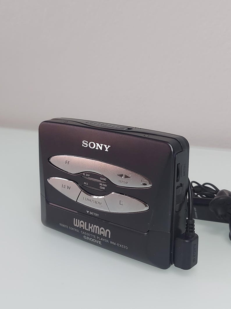 May include: A black Sony Walkman cassette player with silver buttons and the text "WALKMAN" and "GROOVE". The player has a remote control and a battery indicator. The device is a vintage audio player.