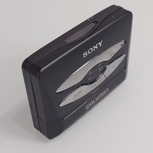 May include: A black Sony Walkman cassette player. The device features silver buttons for rewind, fast forward, and other functions. The word "WALKMAN" is printed on the front, along with the Sony logo. The device is rectangular and has a sleek, retro design.