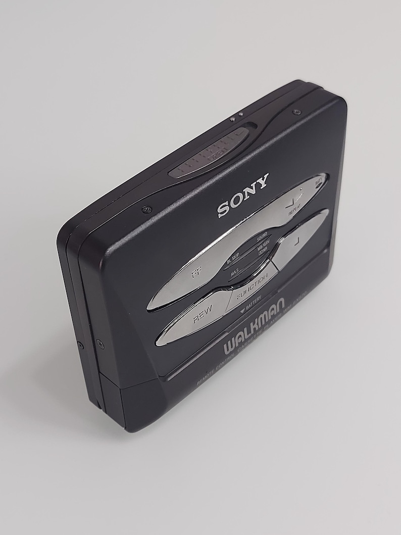 May include: A black Sony Walkman cassette player. The device features silver buttons for rewind, fast forward, and other functions. The word "WALKMAN" is printed on the front, along with the Sony logo. The device is rectangular and has a sleek, retro design.