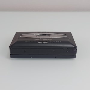 May include: Black Sony Walkman WM-EX570 remote control cassette player. It features silver function buttons and the word "Groove". The device has a rectangular shape and a sleek design.