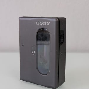 May include: A grey Sony Walkman cassette player. The device is rectangular with rounded corners. The word "SONY" is printed in white on the top. A clear window reveals the cassette tape compartment. A directional arrow is visible.