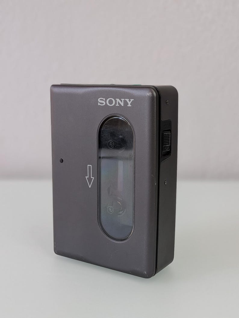 May include: A grey Sony Walkman cassette player. The device is rectangular with rounded corners. The word "SONY" is printed in white on the top. A clear window reveals the cassette tape compartment. A directional arrow is visible.
