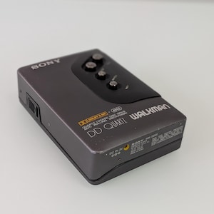 May include: A grey Sony Walkman cassette player. The device has a black top with three buttons and the text "SONY" on the top left. The text "DD QUARTZ WALKMAN" is visible on the front. The device has "DOLBY B NR" and "DC IN 3V" labels.