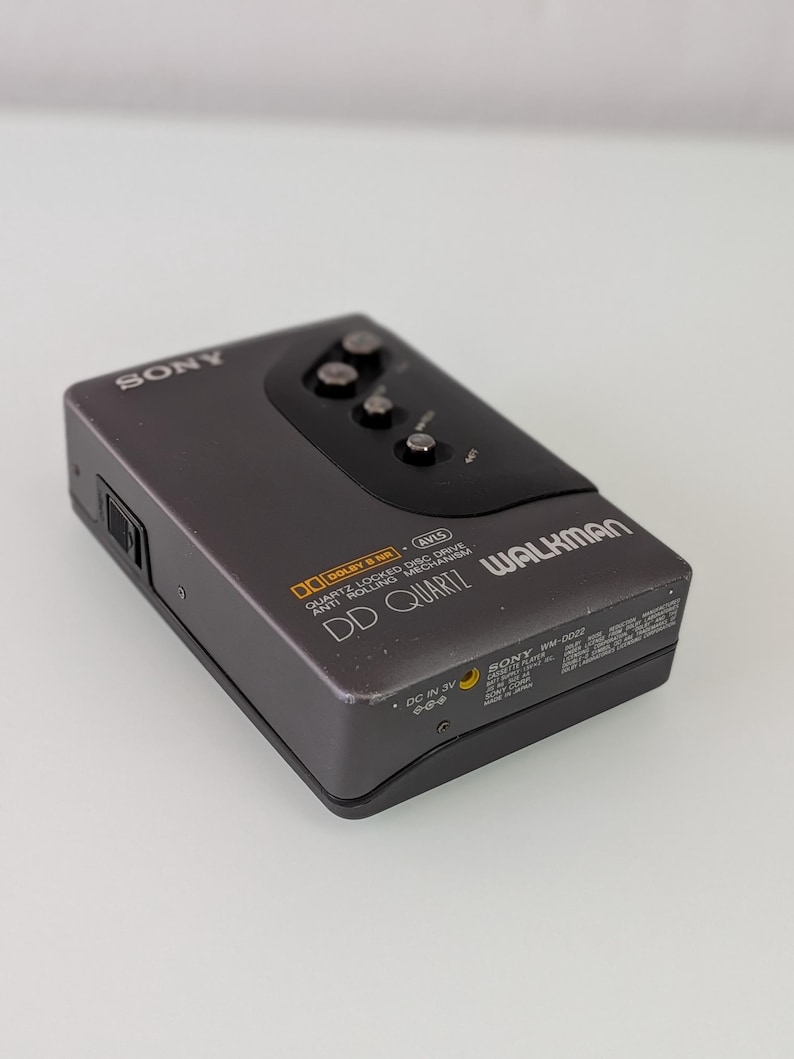 May include: A grey Sony Walkman cassette player. The device has a black top with three buttons and the text "SONY" on the top left. The text "DD QUARTZ WALKMAN" is visible on the front. The device has "DOLBY B NR" and "DC IN 3V" labels.