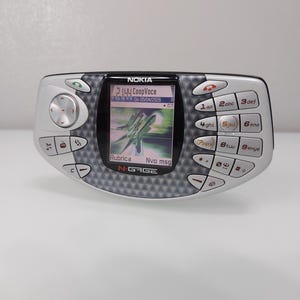Può includere: Dispositivo di gioco portatile Nokia N-Gage argentato. Il dispositivo ha uno schermo nero che mostra un'immagine di gioco e testo. È dotato di tastierino, pad direzionale e vari pulsanti funzione. Il logo Nokia è visibile in alto.