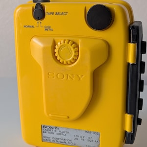 May include: A bright yellow Sony WM-B53 portable cassette player featuring a black volume dial, a tape select switch for normal, CrO2, and metal tapes, and a central locking mechanism. The device includes a label detailing battery requirements.