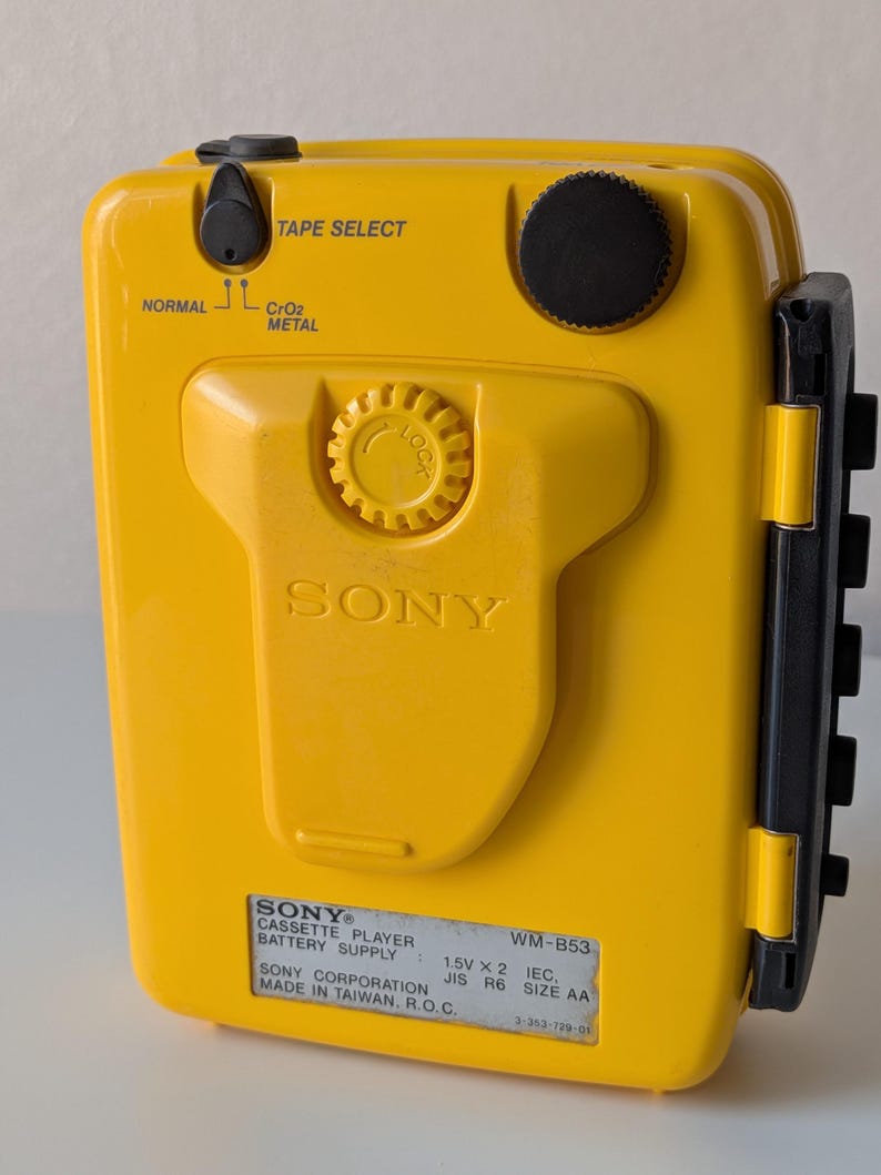 May include: A bright yellow Sony WM-B53 portable cassette player featuring a black volume dial, a tape select switch for normal, CrO2, and metal tapes, and a central locking mechanism. The device includes a label detailing battery requirements.