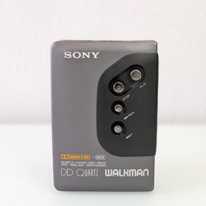 May include: A grey Sony Walkman DD Quartz portable cassette player. The device has a black control panel with play, stop, rewind, and fast forward buttons. The text "SONY" and "WALKMAN" are visible on the device.