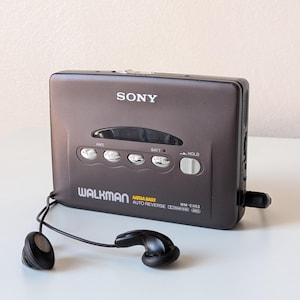 May include: A dark gray Sony Walkman cassette player with a rectangular body, featuring the text SONY, WALKMAN, MEGA BASS, AUTO REVERSE, and WM-EX53. The device includes four silver control buttons, a hold switch, and black wired earbuds.