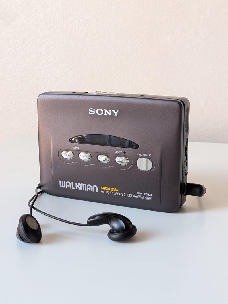 May include: A dark gray Sony Walkman cassette player with a rectangular body, featuring the text SONY, WALKMAN, MEGA BASS, AUTO REVERSE, and WM-EX53. The device includes four silver control buttons, a hold switch, and black wired earbuds.