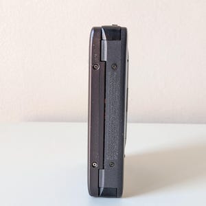 May include: A dark gray electronic device with a rectangular, elongated body. The side profile shows a textured surface with two visible metal screws and small, embossed text detailing manufacturing information and licensing specifications.